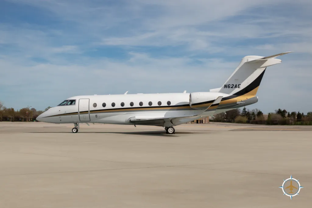 A white business jet with gold and black stripes on the tail, registration N62AE, parked on a tarmac under a blue sky.