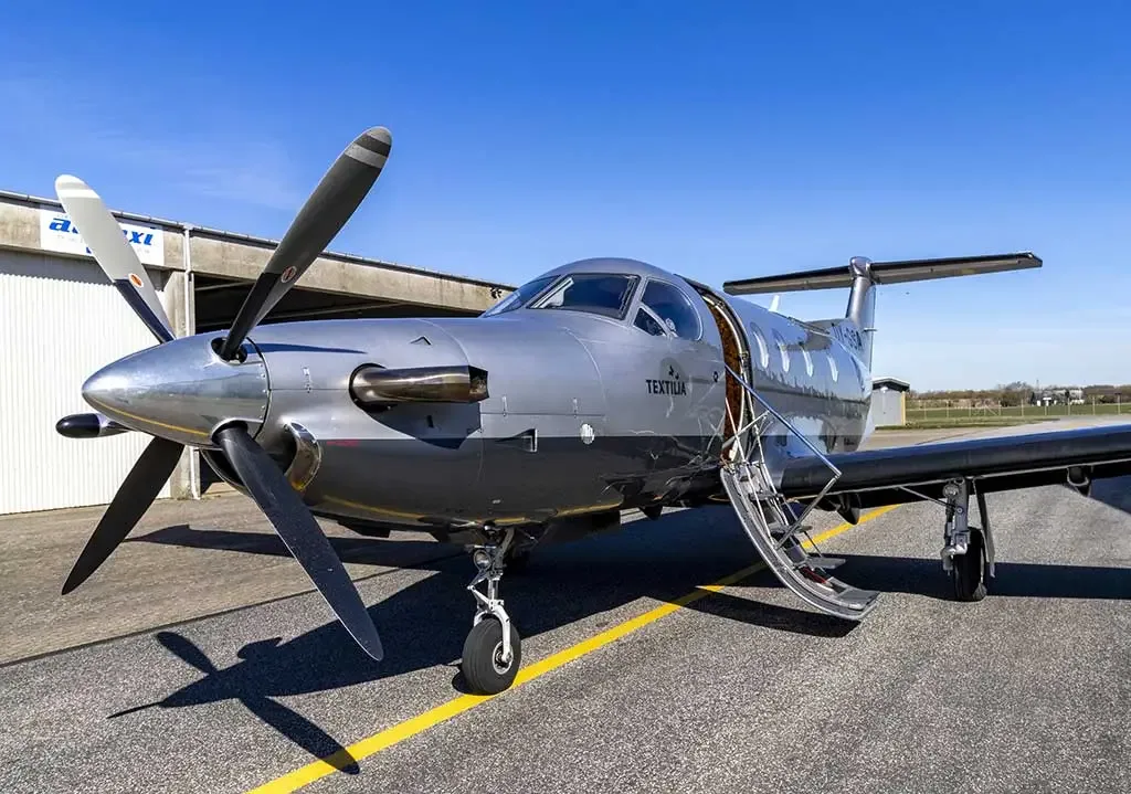 A silver and dark grey Pilatus PC-12 aircraft with its passenger door open, parked on an asphalt tarmac.