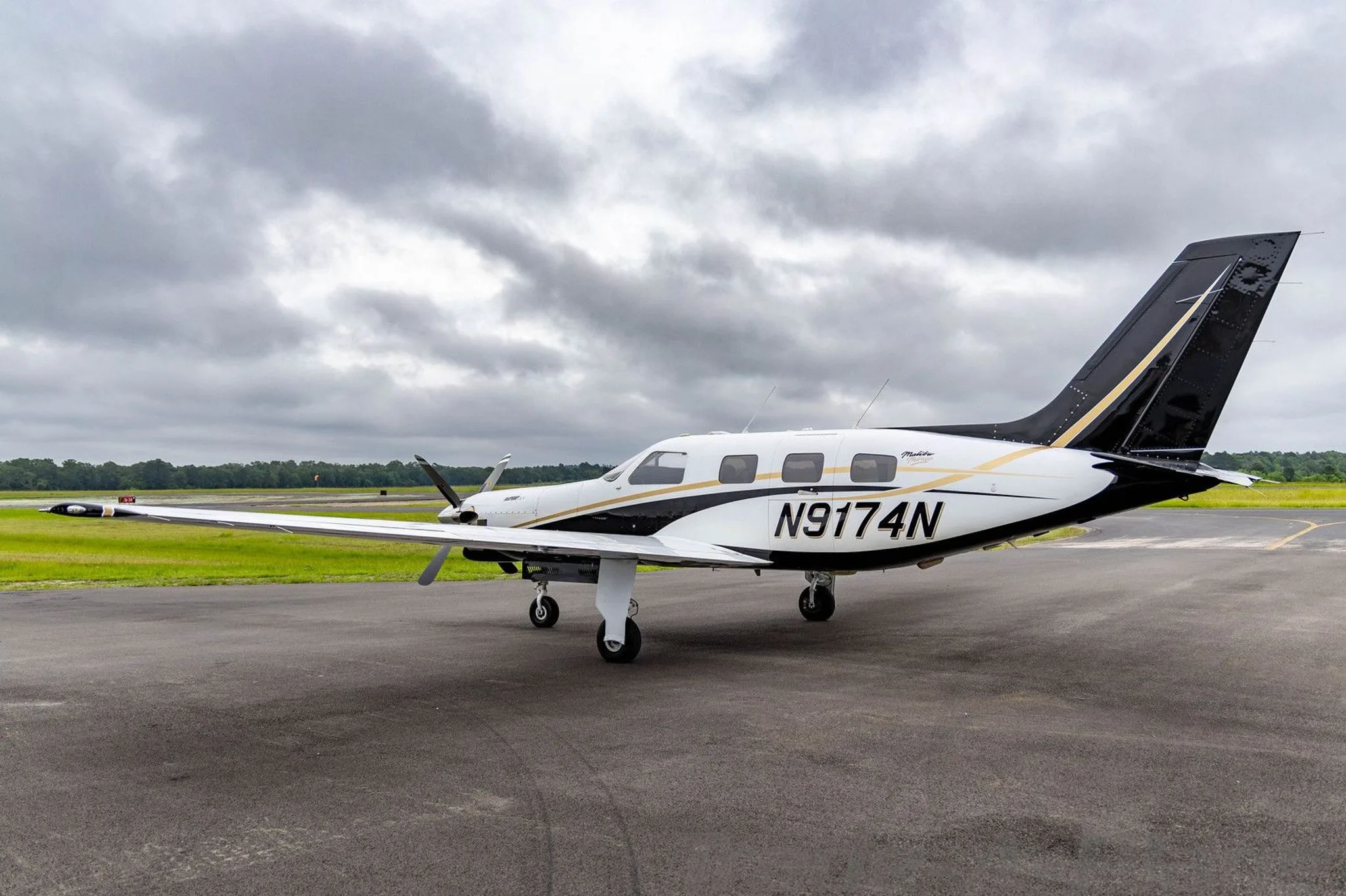 A white and black Piper Malibu Mirage (JetPROP DLX) N9174N with gold accents parked on a tarmac, viewed from the rear-left.