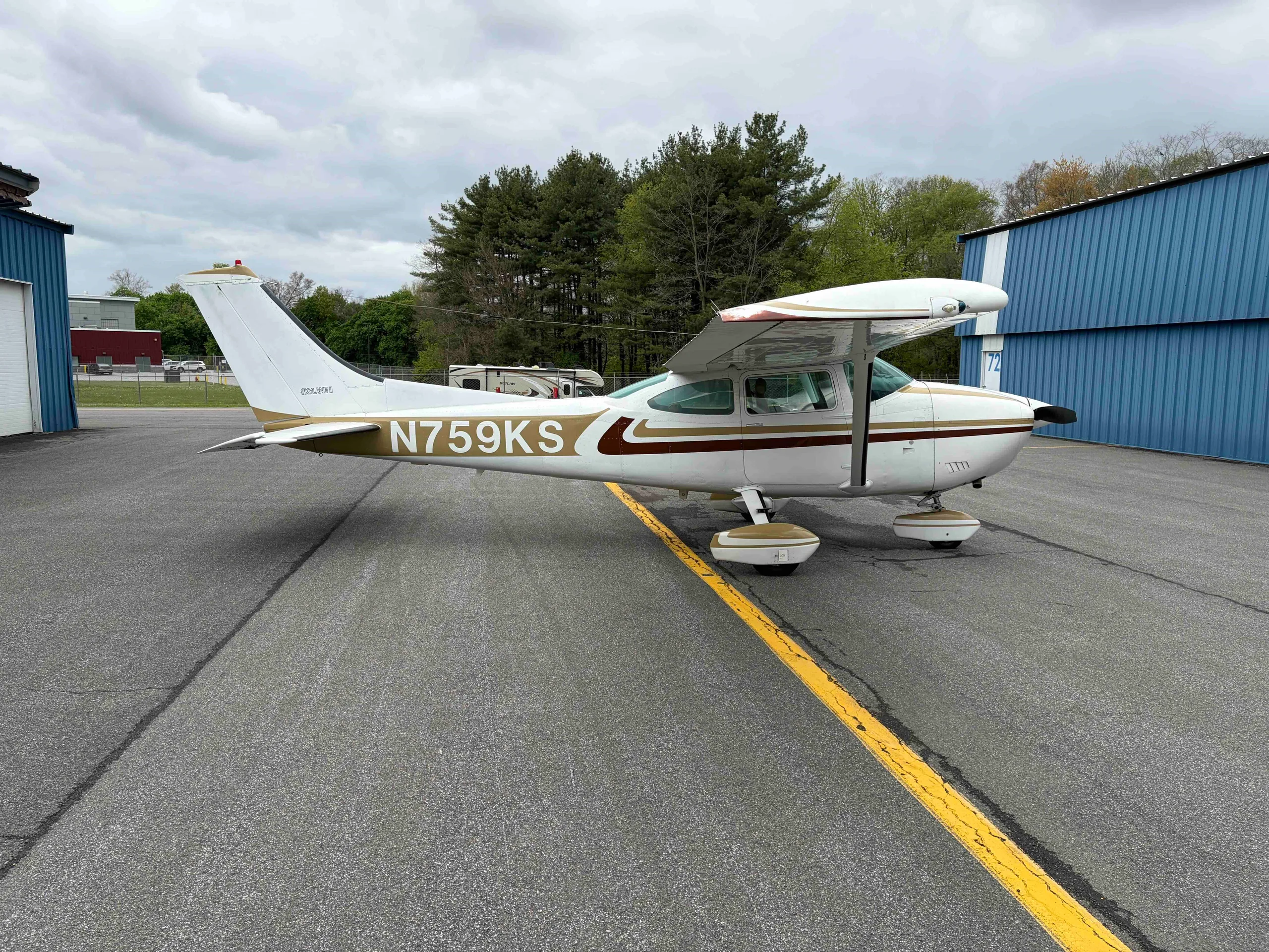 A three-quarter rear view of the white Cessna 182 Skylane N759KS with gold and brown stripes, parked on an airport tarmac.
