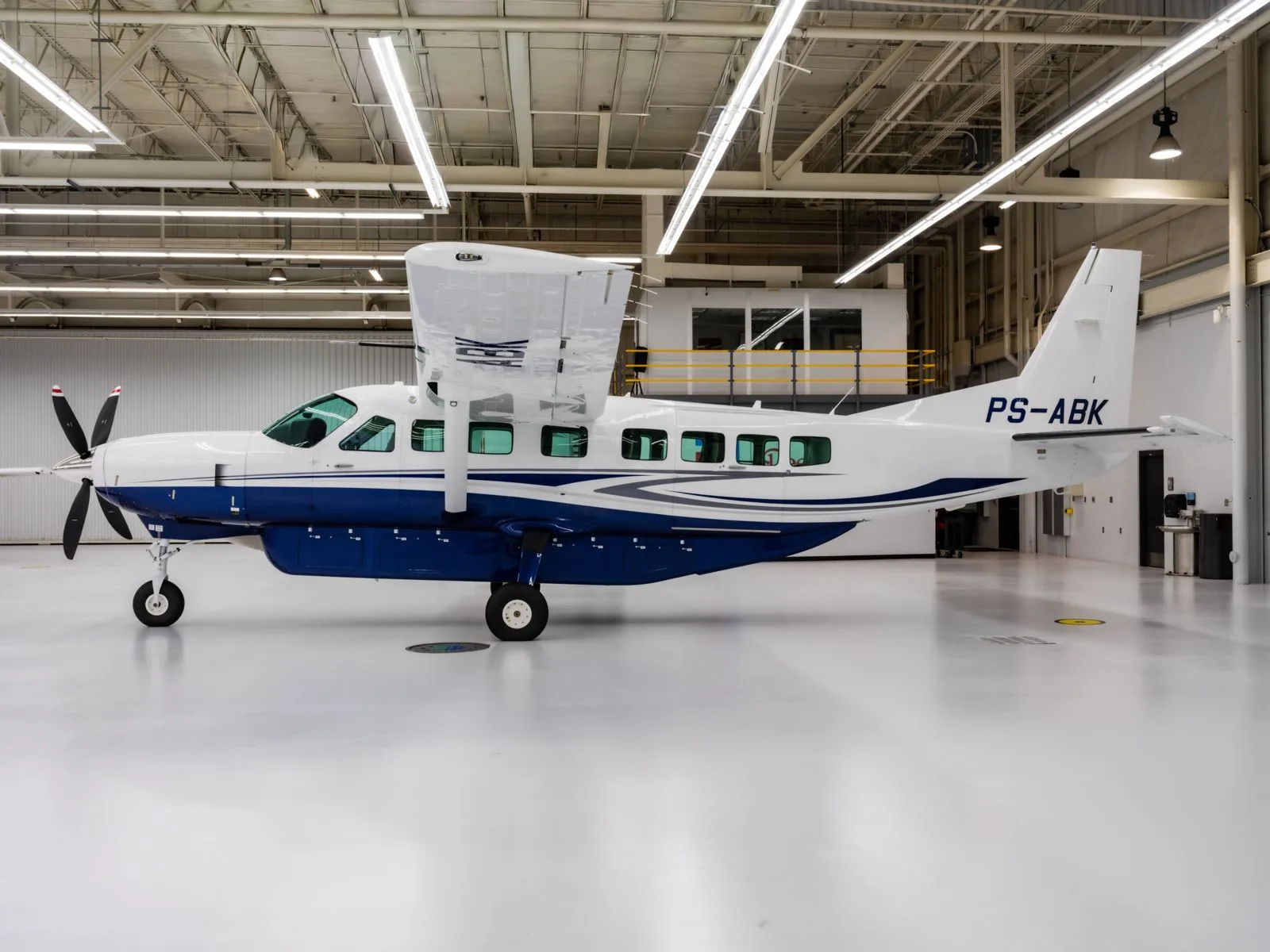 A white and blue Cessna 208B Grand Caravan (PS-ABK) is parked inside a brightly lit hangar, viewed from its left side.