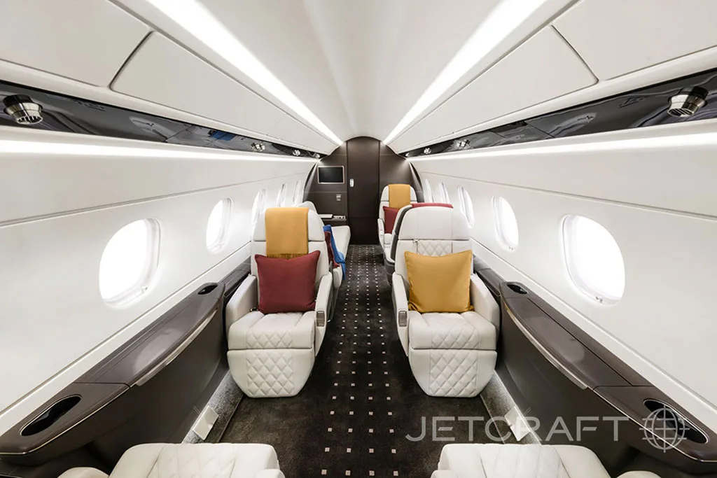 The luxurious interior cabin of a business jet with white quilted seats, dark carpet, and yellow and red accent pillows.