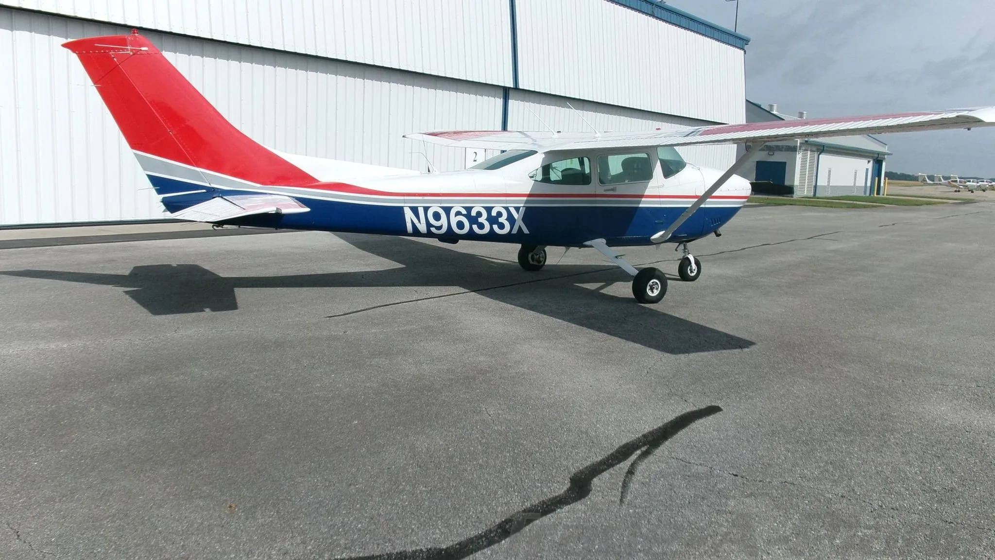 A three-quarter rear view of a white, blue, and red Cessna 182 Skylane with registration N9633X parked on an asphalt apron.