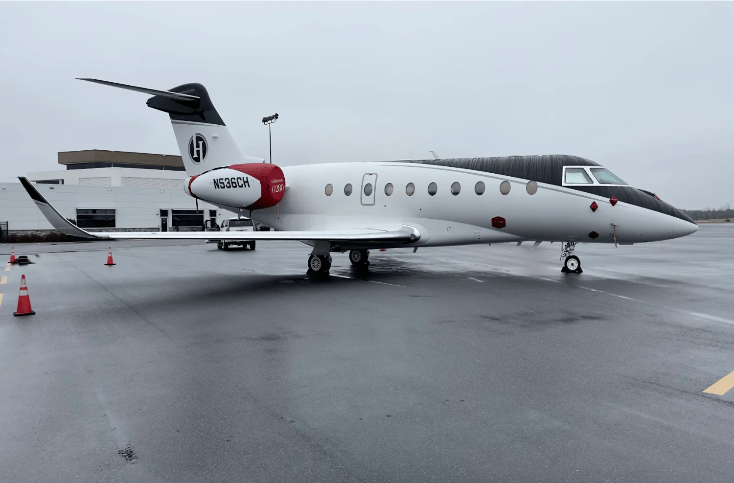 A white and black Gulfstream G280 business jet with red accents on the tail and engine nacelle, parked on a wet tarmac.