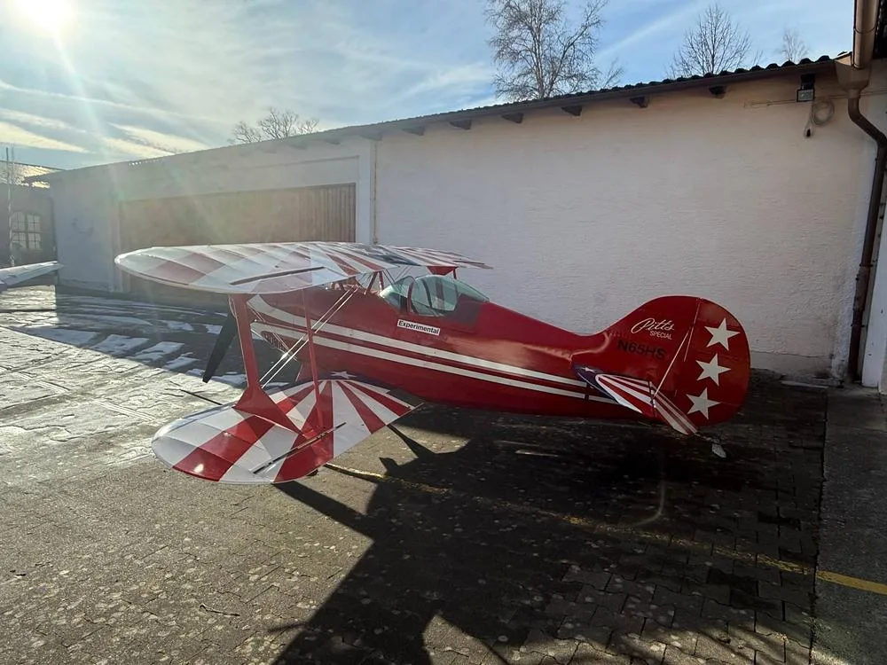A red Pitts Special biplane with white stripes on the wings and tail, parked on an asphalt surface next to a white building under a sunny sky.