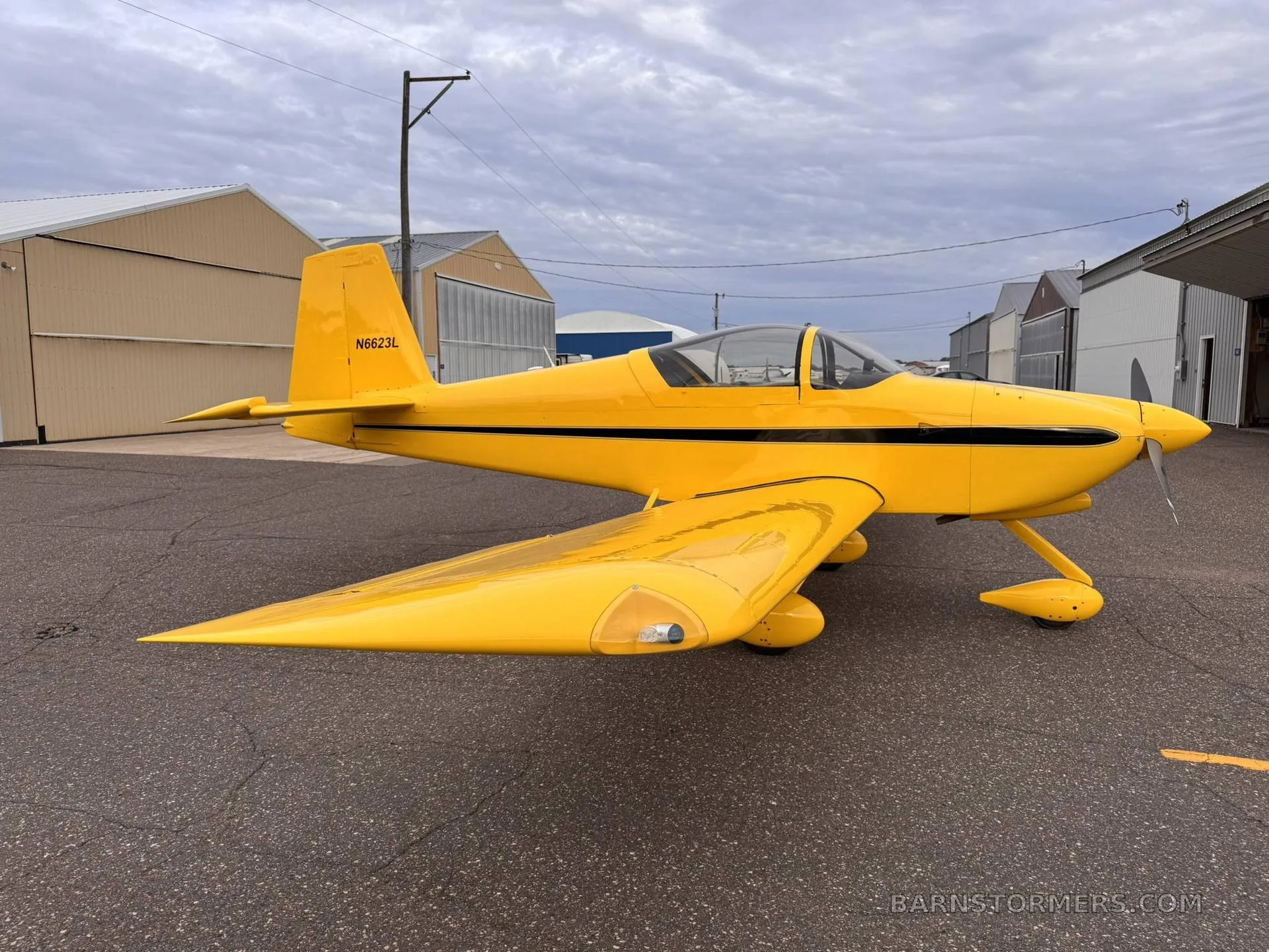 A bright yellow single-engine aircraft with a black stripe and registration N6623L is parked on an asphalt ramp with hangars in the background.