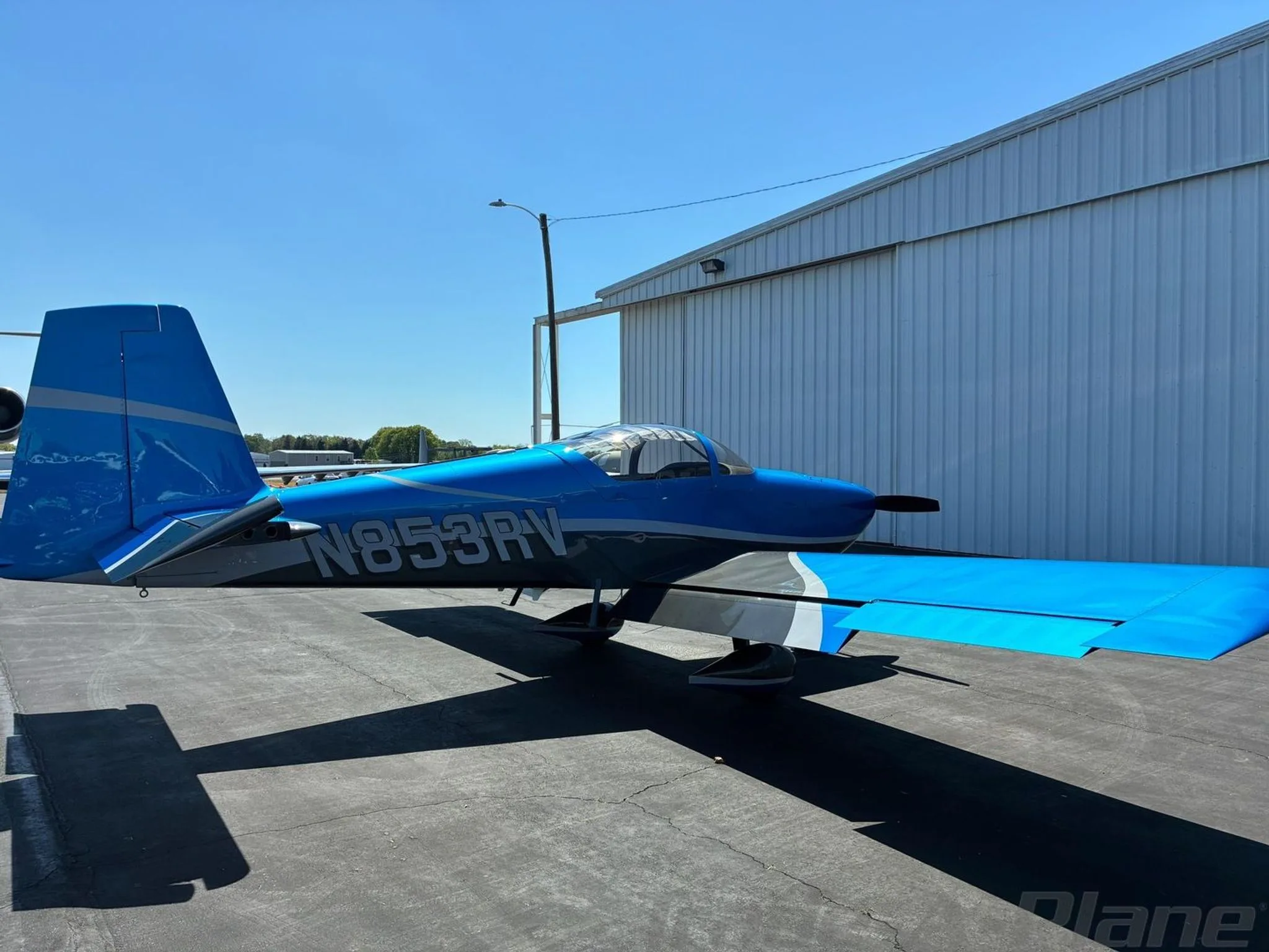 A blue and grey N853RV aircraft parked on an airfield with its wing extended, next to a white hangar.