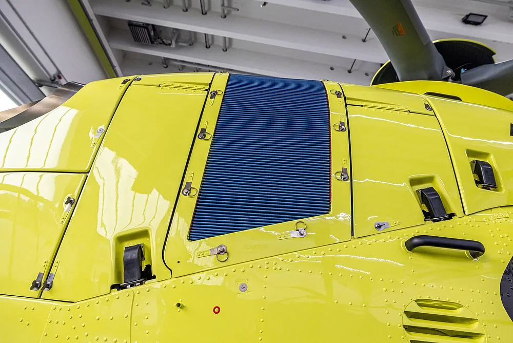 A close-up view of the bright yellow fuselage of an Airbus H135 helicopter, showing a blue engine vent.