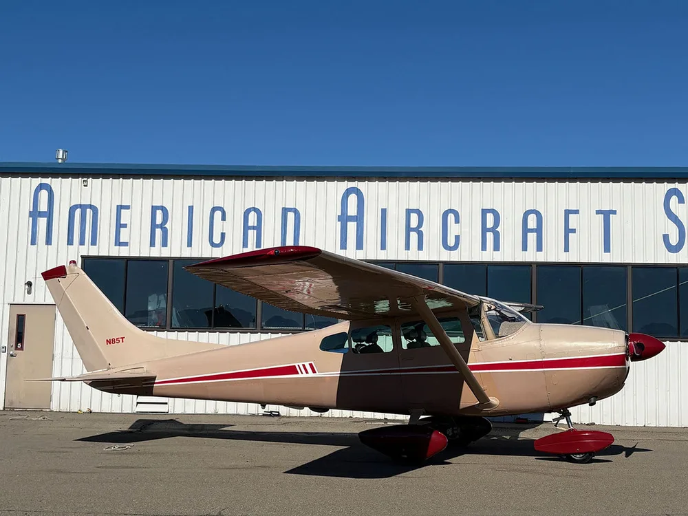 A beige and red Cessna 182 aircraft, registration N85T, parked with its nose facing right, in front of a hangar.