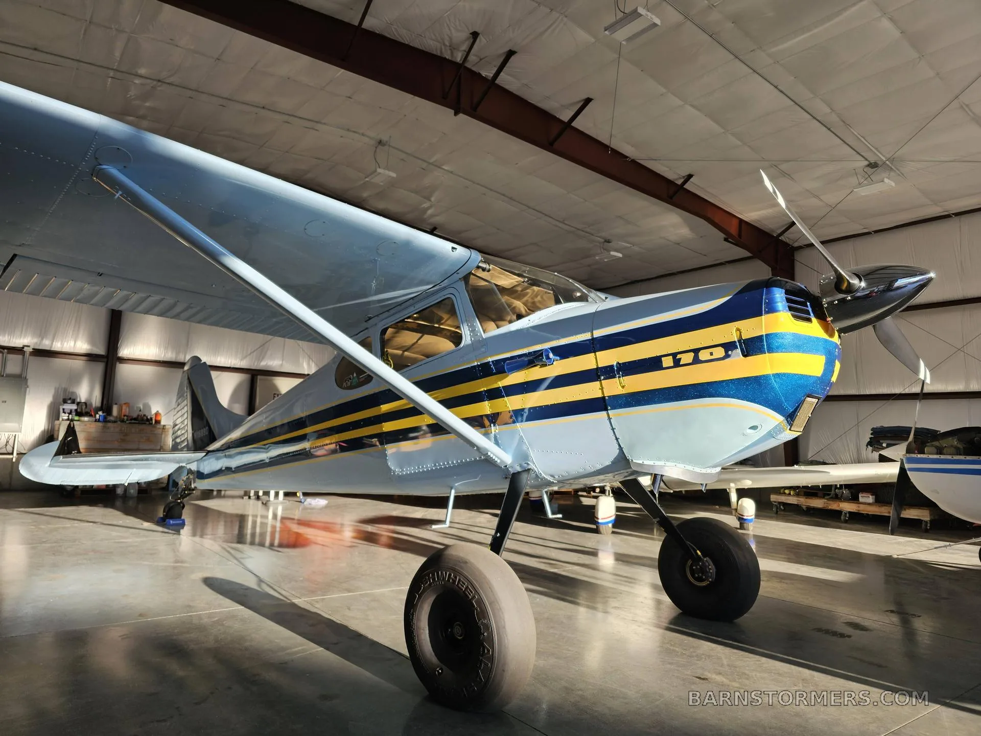 A light blue and yellow striped Cessna 170 aircraft is parked inside a hangar.