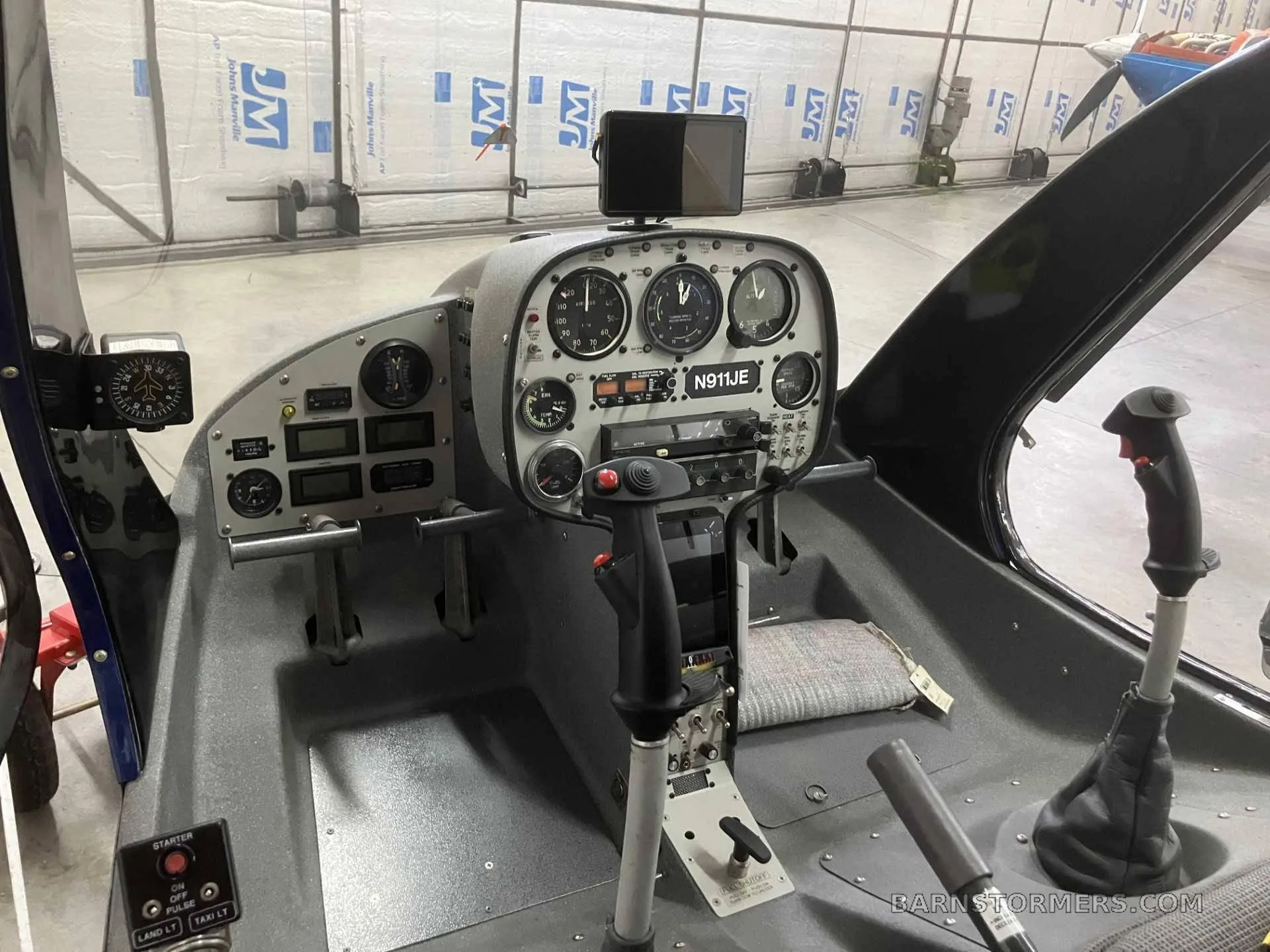 The cockpit of a helicopter (N911JE) showing the instrument panel, cyclic, and collective controls inside a hangar.