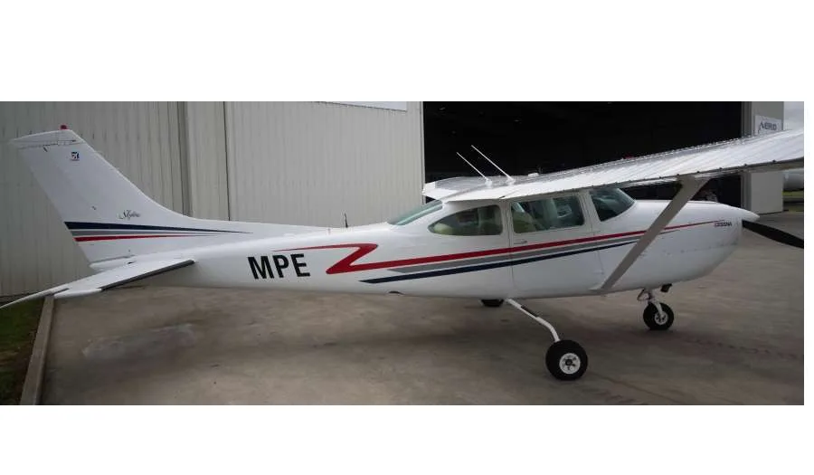 A white Cessna aircraft with red and blue stripes and 'MPE' on the tail, parked in front of a hangar.