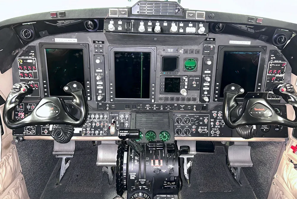 The cockpit instrument panel of an aircraft features multiple digital displays, gauges, and two control yokes.