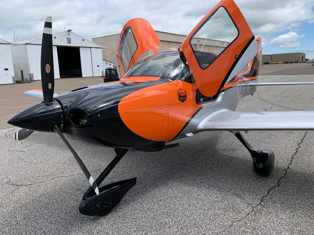 Front view of a Cirrus SR20 aircraft with orange, black, and silver paint scheme, doors open, on an airfield.