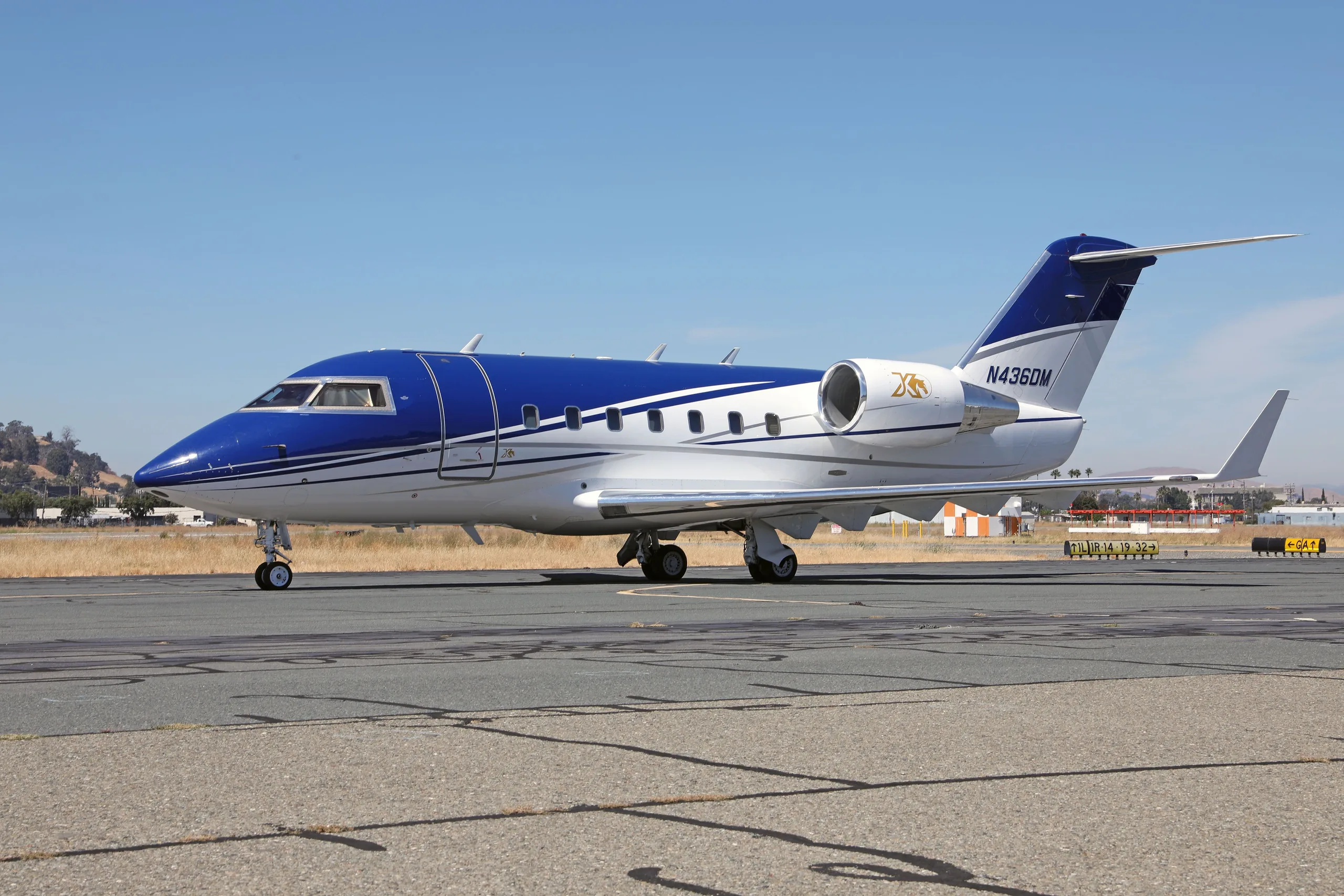 A blue and white Bombardier Challenger 604 aircraft (N436DM) parked on a tarmac under a clear sky.