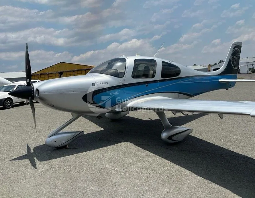 A silver Cirrus SR22 aircraft with blue accents on the fuselage, parked on an asphalt ramp under a partly cloudy sky.