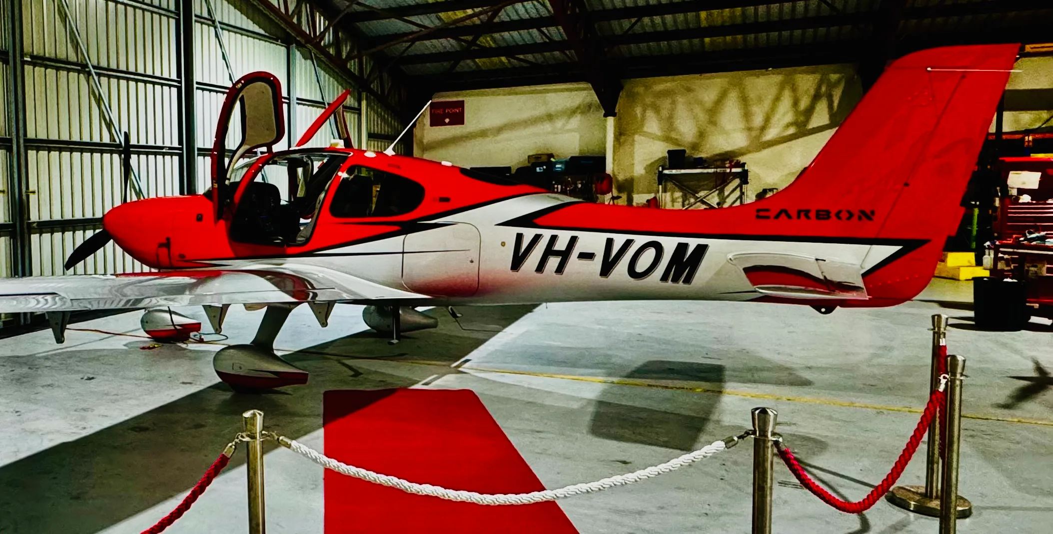 A red and white single-engine propeller aircraft, registration VH-VOM, with its doors open, parked inside a hangar.