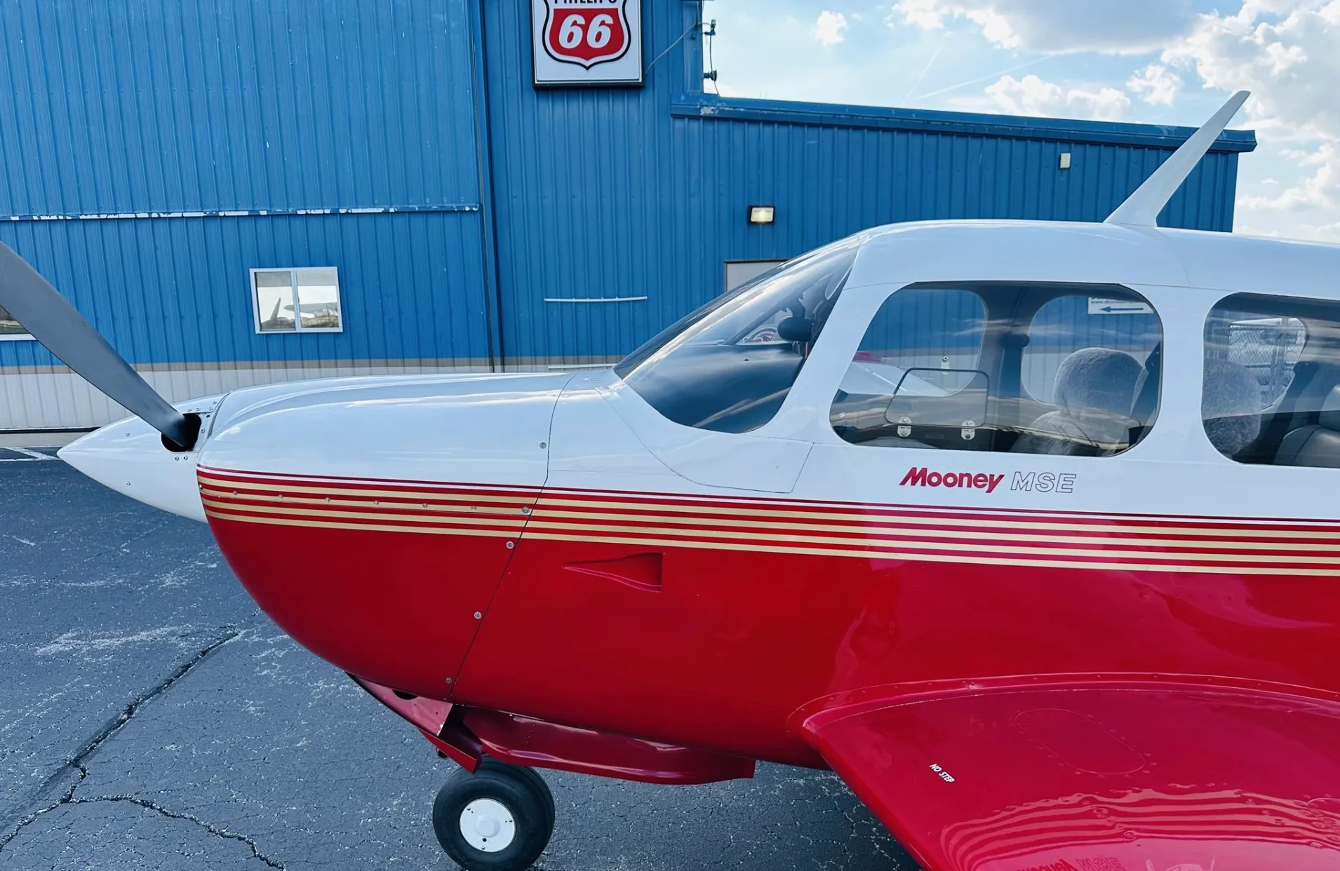 A close-up of the front right side of a red and white Mooney MSE aircraft parked in front of a blue hangar.