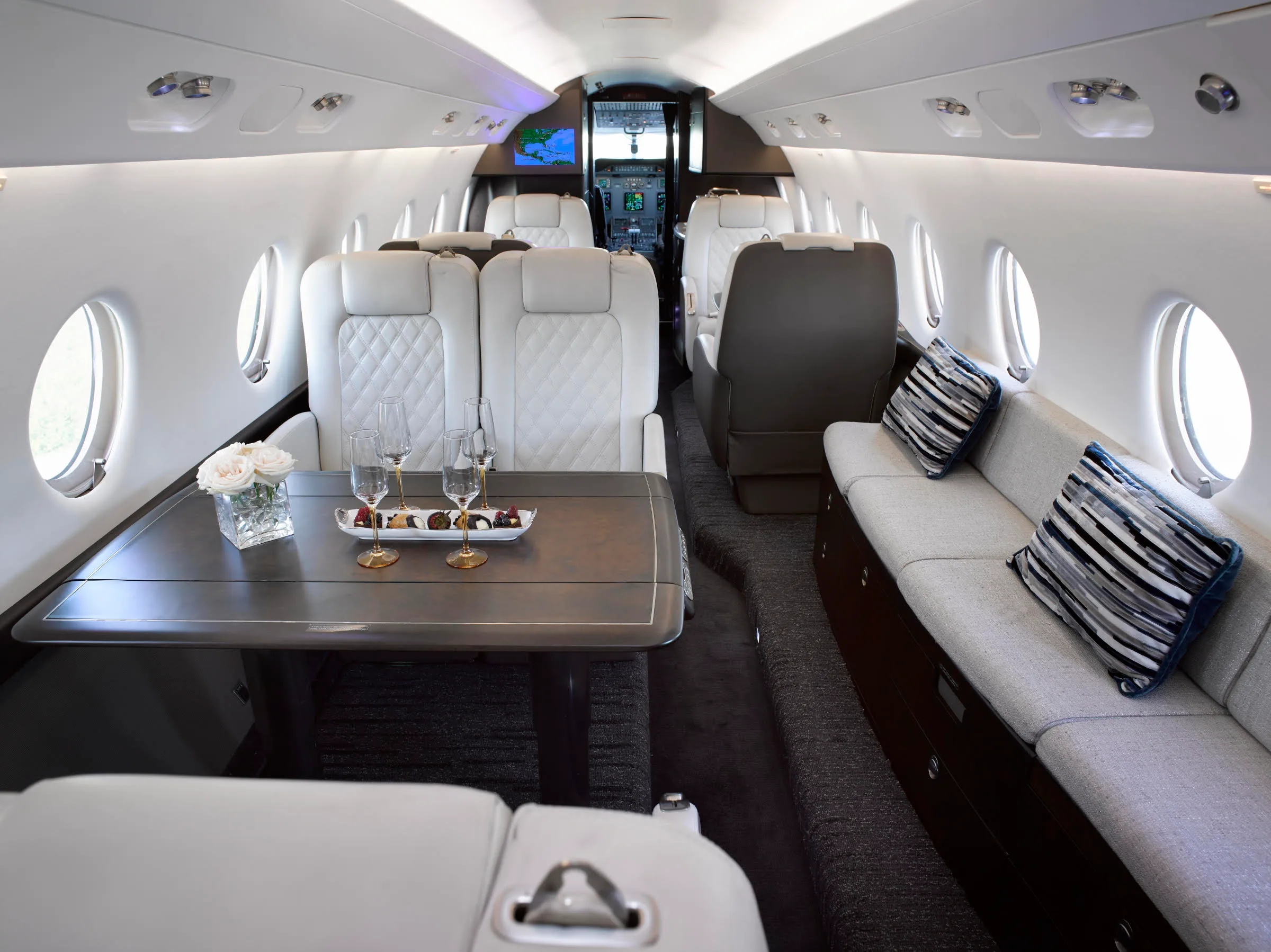 The luxurious interior of a private jet cabin features white quilted seats, a table set with drinks and snacks, and a view towards the cockpit.