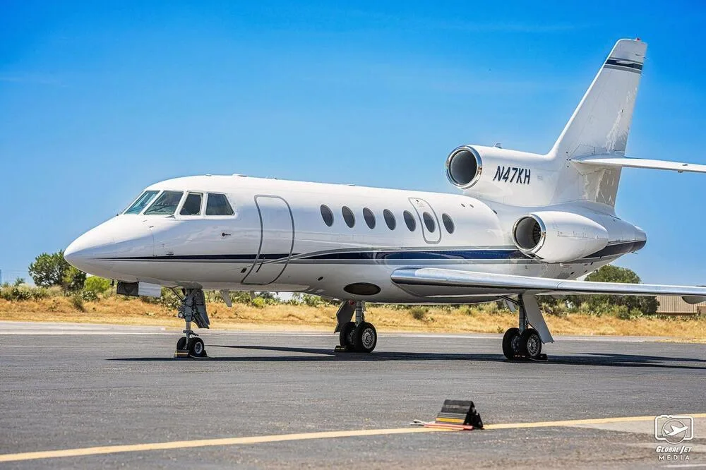 A side view of a white Dassault Falcon 900EX business jet with blue stripes, parked on an airfield.