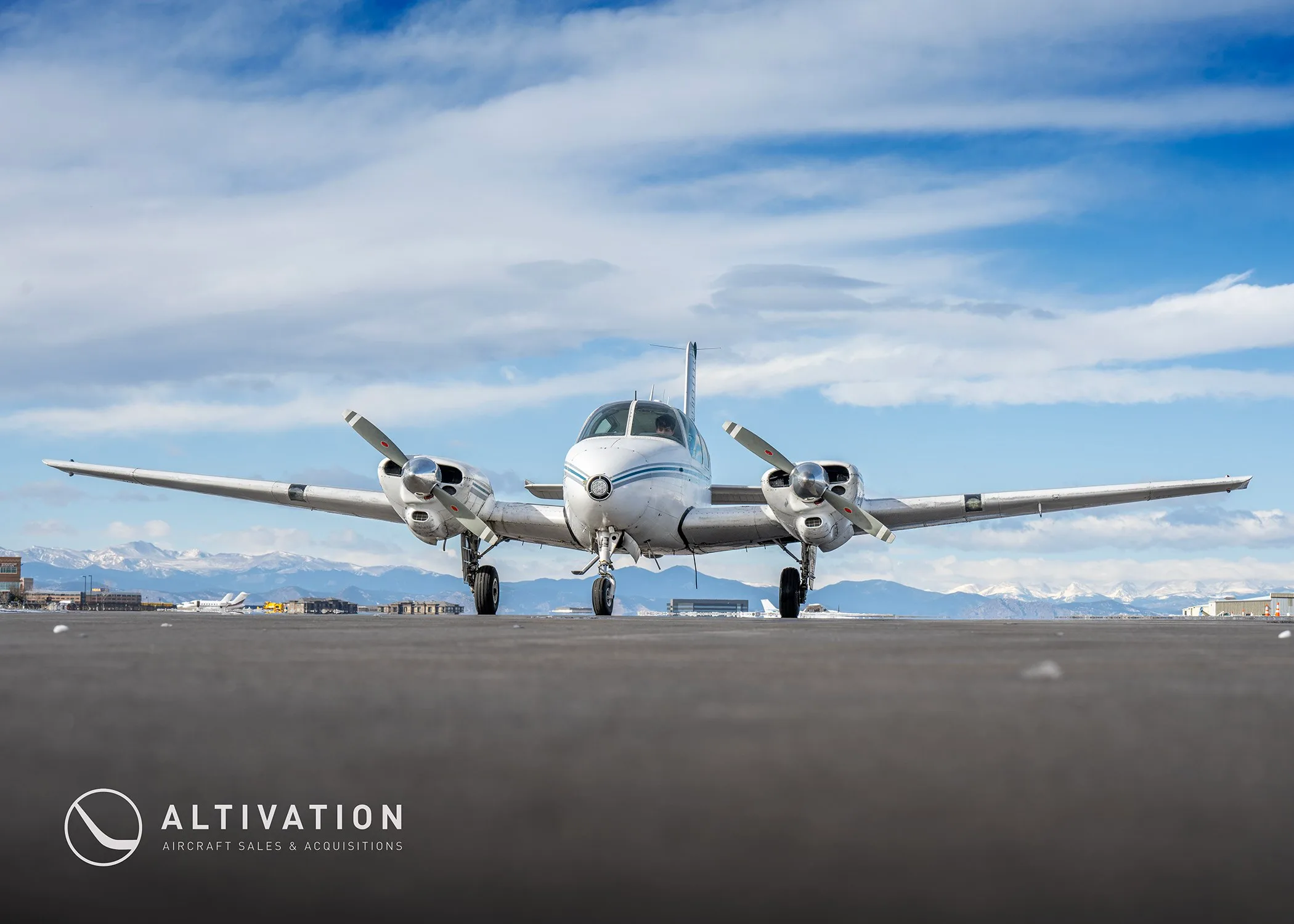 Front view of a white and blue twin-engine propeller aircraft on a tarmac with mountains in the background.