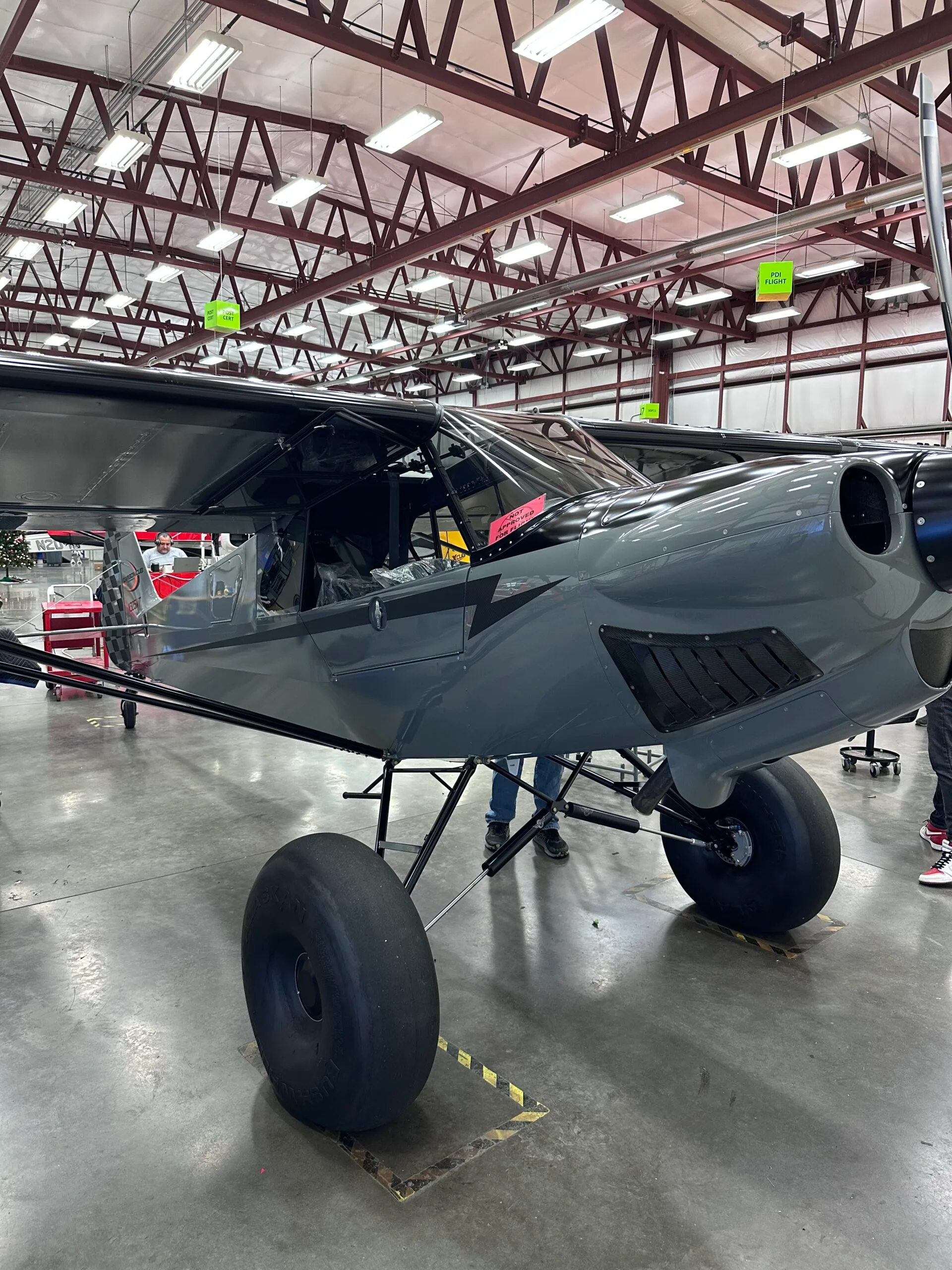A grey bush plane with large tires is parked inside a hangar, viewed from the front-right side.