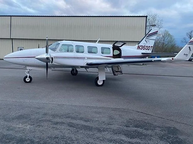 A white twin-engine propeller aircraft, N350DT, with dark stripes is parked on an asphalt ramp in front of a hangar with its passenger door open.