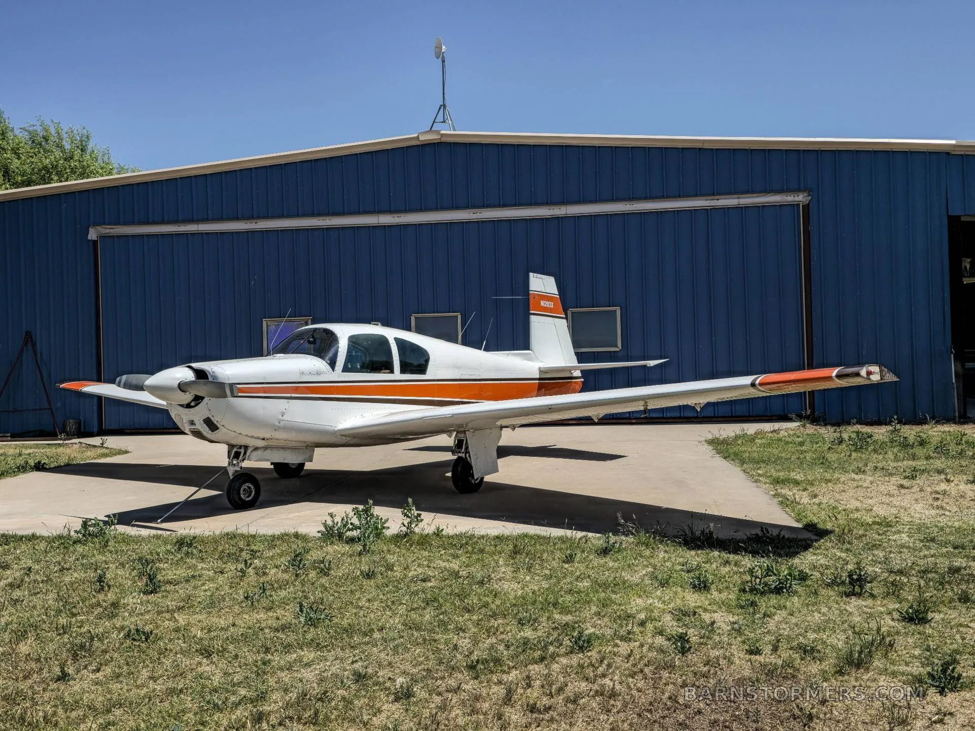 A white and orange single-engine aircraft (N282X) parked on a concrete pad and grass in front of a blue hangar.