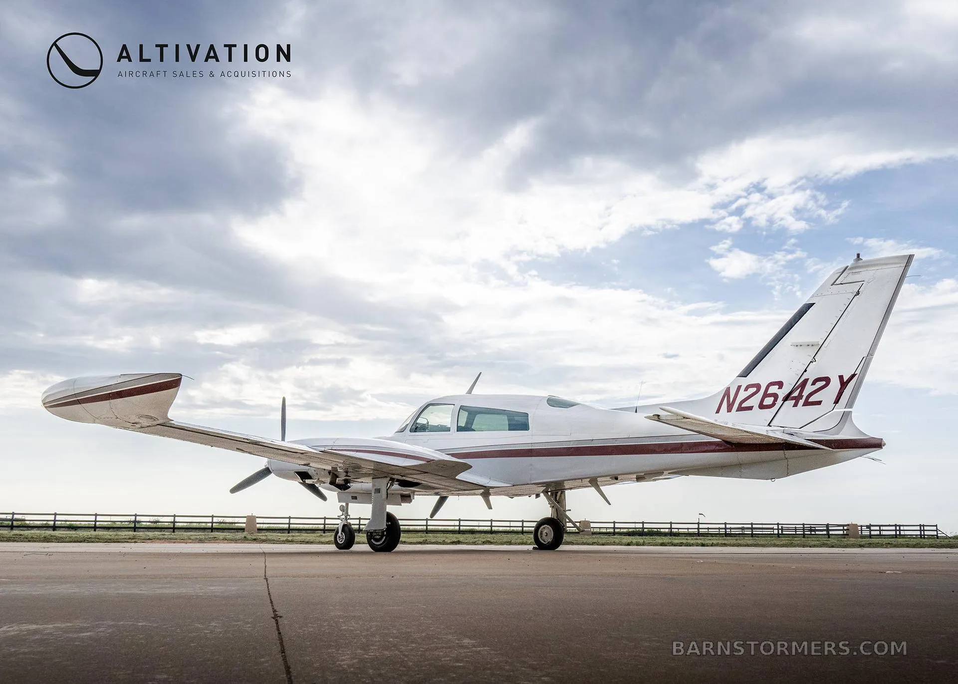A white twin-engine propeller aircraft with maroon stripes, registration N2642Y, viewed from the rear-side on an asphalt runway under a cloudy sky.