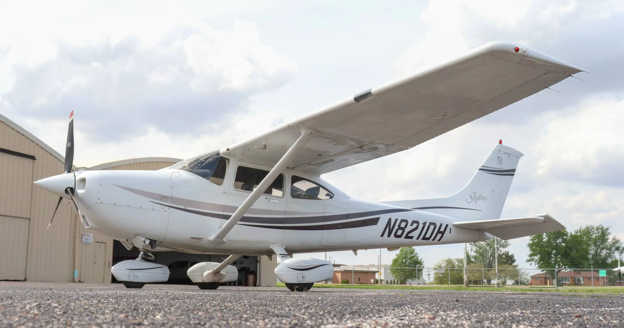 A low-angle side view of the white Cessna Skylane N821DH with dark grey and brown stripes, parked on an asphalt tarmac.