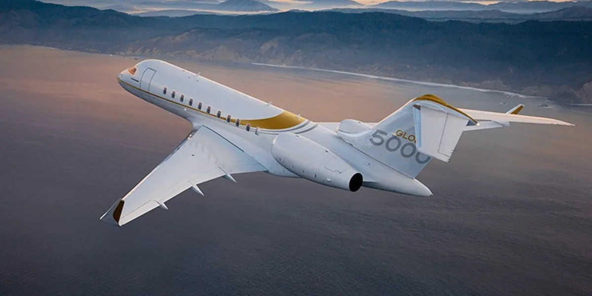 A white and gold Bombardier Global 5000 business jet flies over a body of water with mountains in the background at dusk.