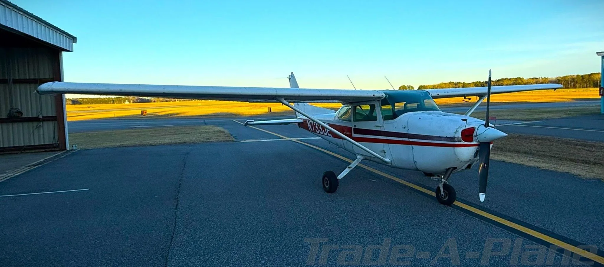 A white Cessna 172N with red and black stripes, registration N733JE, parked on an airfield at sunset.