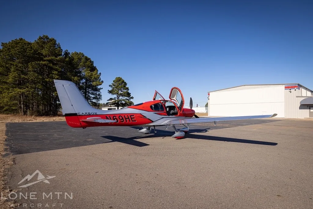 A red and silver Cirrus SR22T aircraft with open doors parked on an asphalt ramp with a hangar in the background.