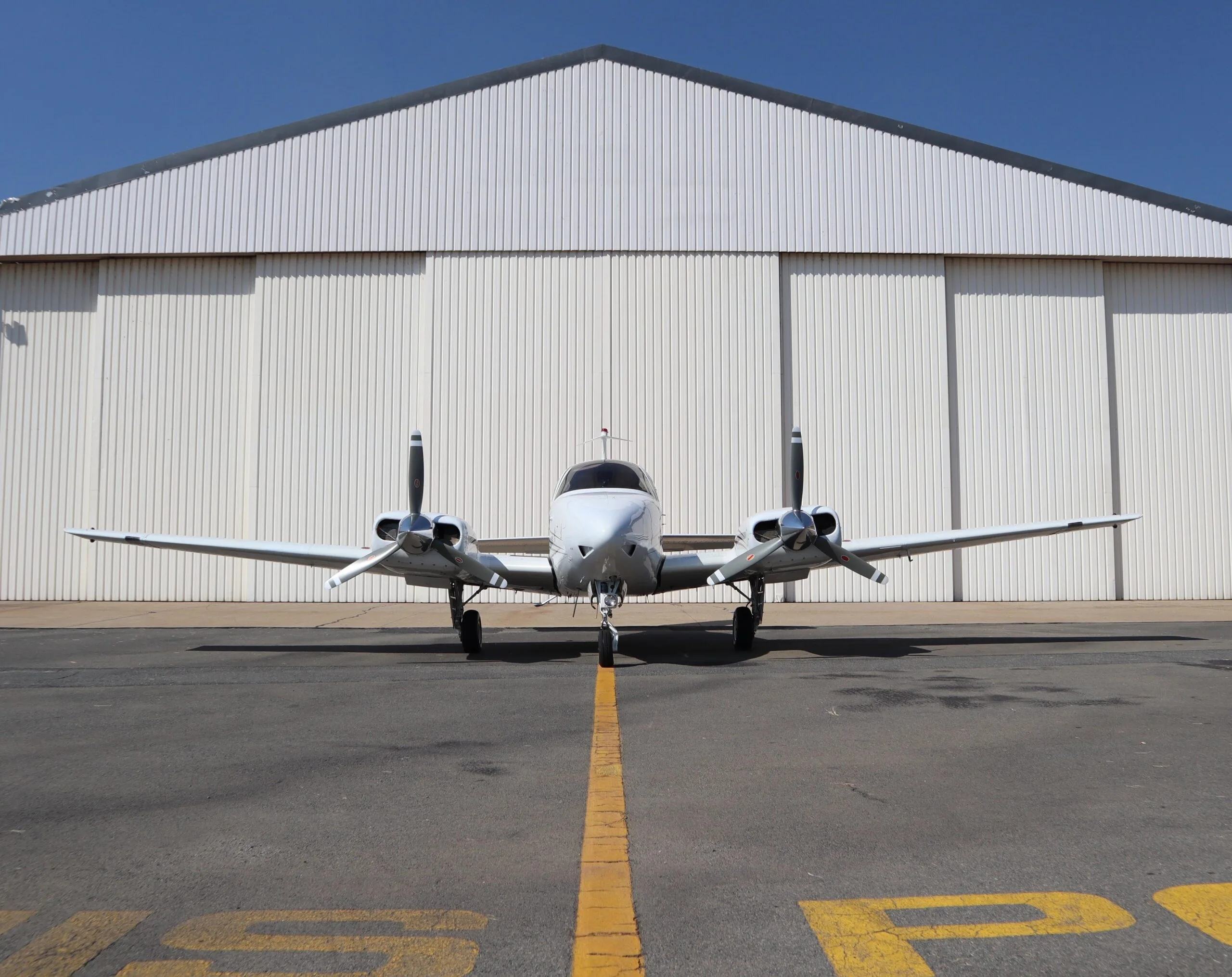 A white twin-engine propeller aircraft is parked on an asphalt tarmac in front of a large white hangar under a clear blue sky.