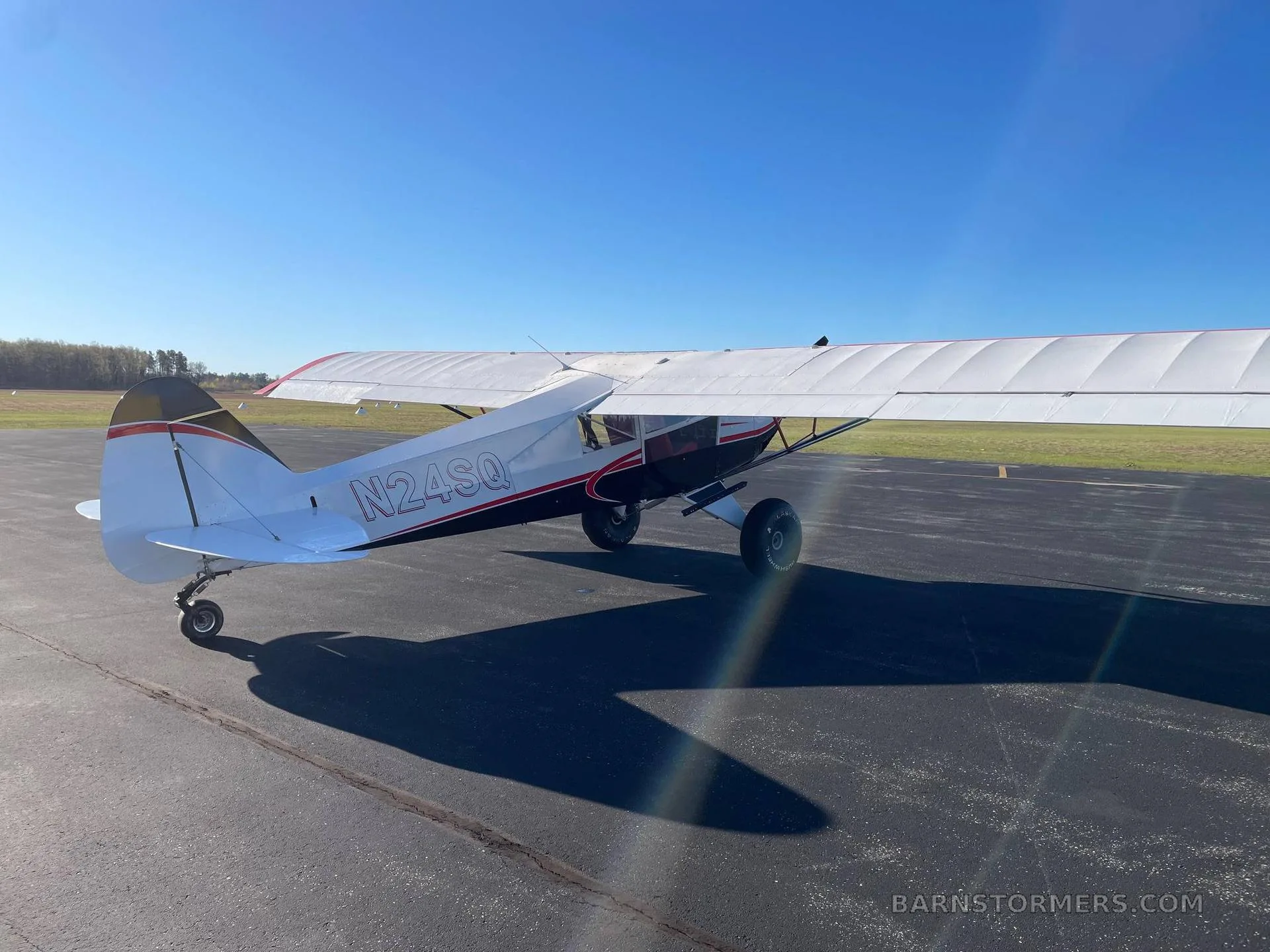 A rear-quarter view of a white and red high-wing aircraft with registration N24SQ, parked on an asphalt apron under a clear blue sky.