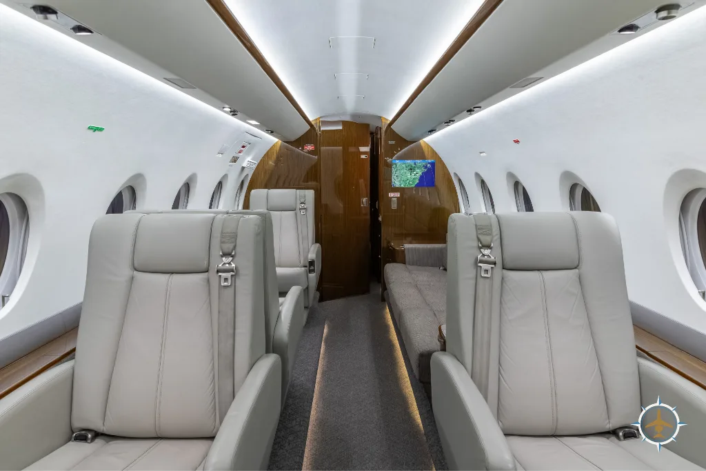 The cabin interior of a business jet, showing light grey seats and a beige sofa on the right side.