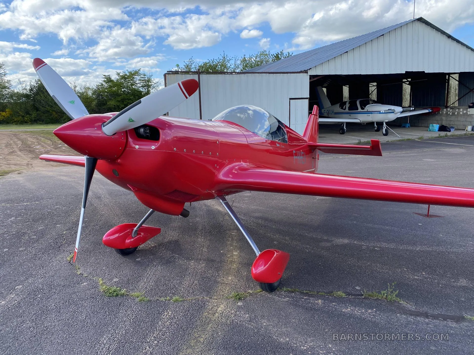 A red single-engine aerobatic aircraft is parked on an asphalt apron in front of an open hangar with another aircraft inside.