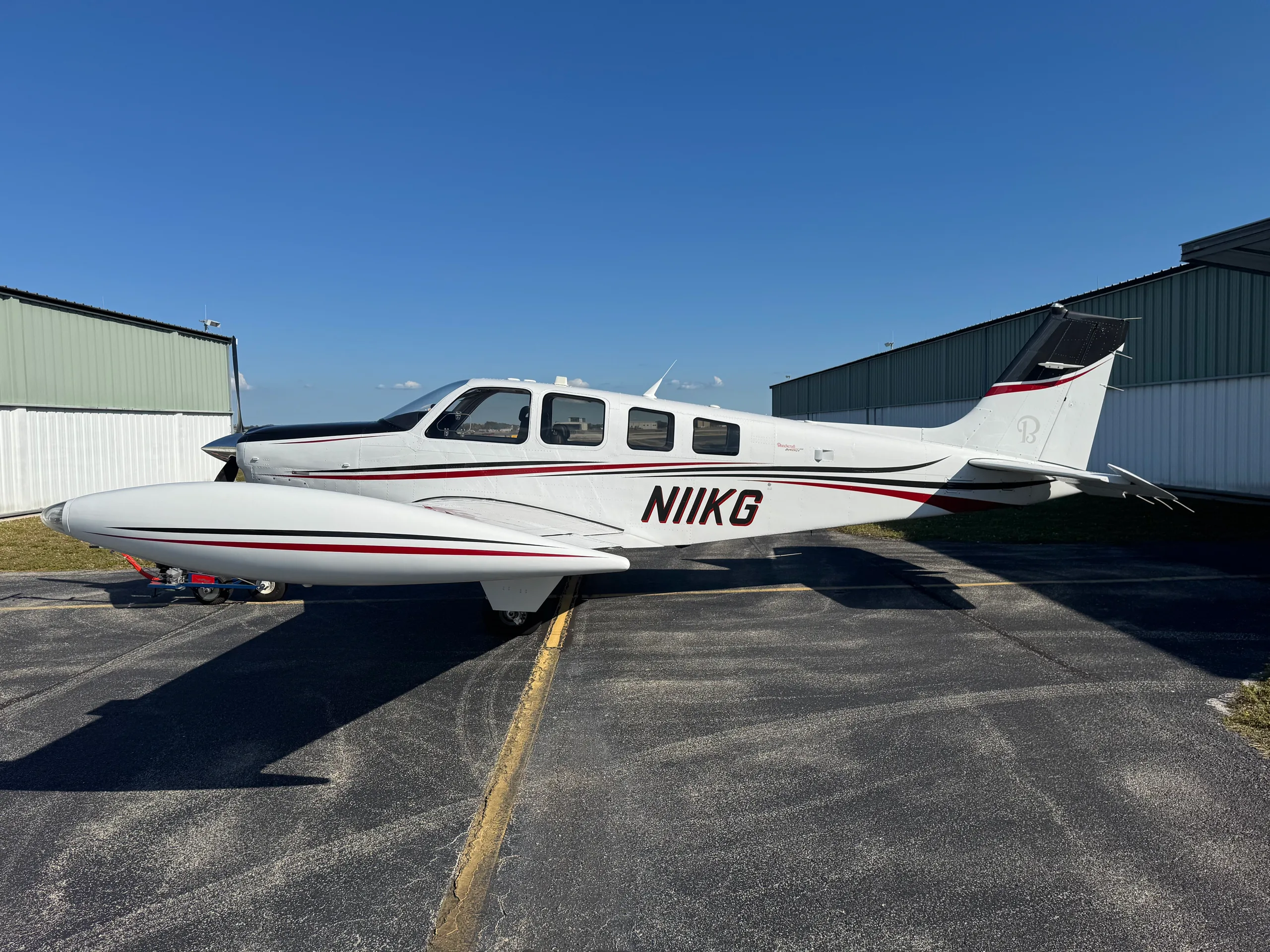 A white Beechcraft Bonanza N11KG with black and red stripes parked on an asphalt ramp next to hangars.