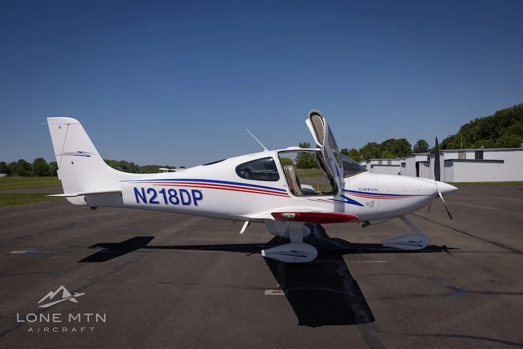 Side view of a white Cirrus aircraft (N218DP) with blue and red stripes and red wingtips, doors open, parked on an airfield.