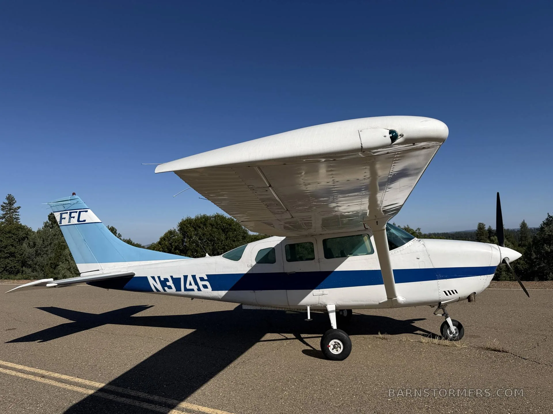 A white and blue Cessna 182 Skylane with registration N3746 parked on an asphalt surface, viewed from the front-right side.