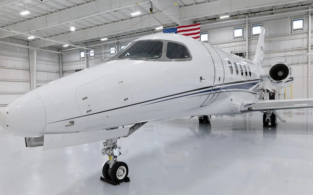 A front-quarter view of a white business jet with blue and black stripes, parked in a hangar with an American flag in the background.