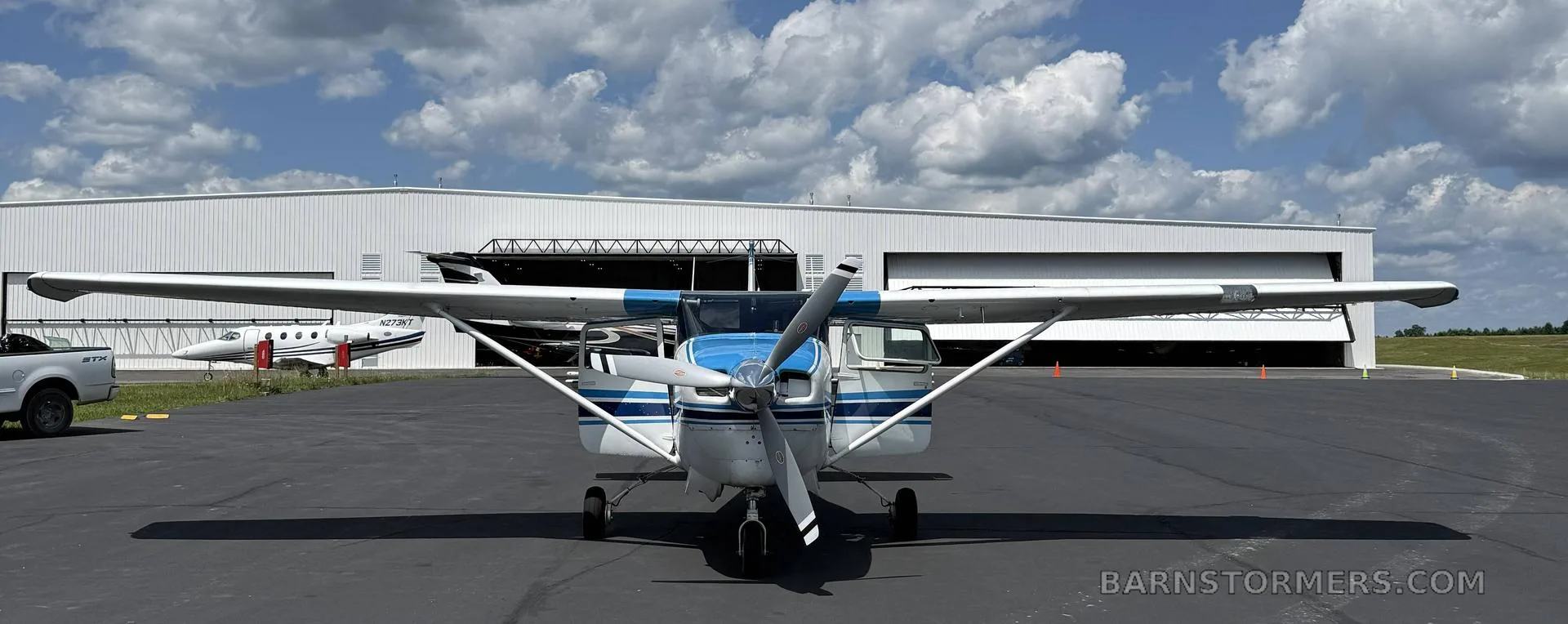 A white and blue Cessna 182 aircraft, N8727Z, is parked on a tarmac in front of hangars under a partly cloudy sky.