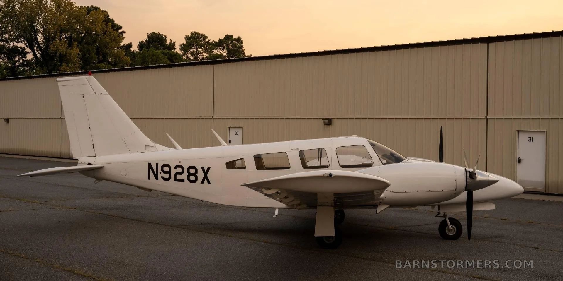 A white twin-engine propeller aircraft (N928X) is parked on an asphalt ramp with a hangar in the background at sunset.
