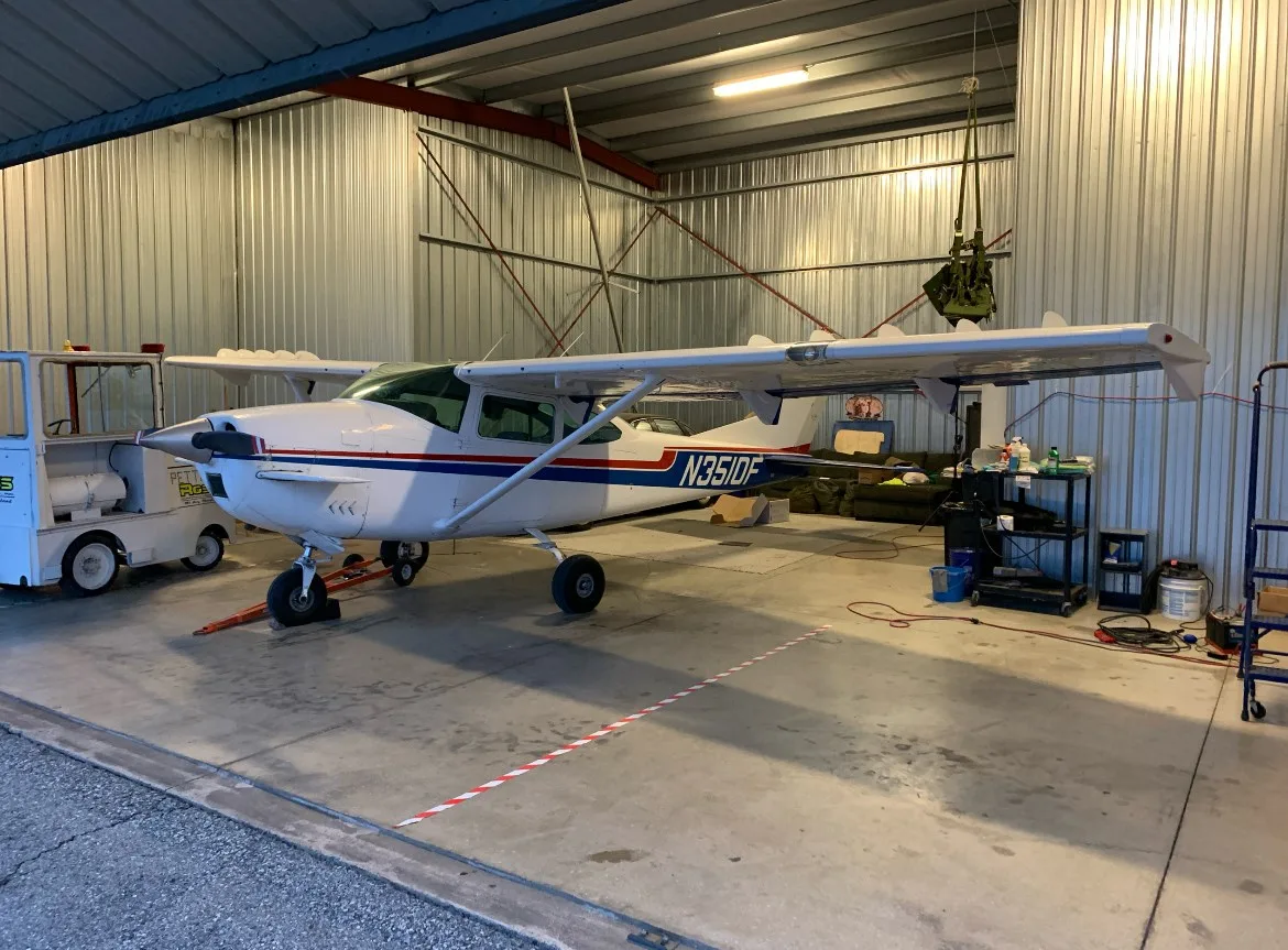 A white Cessna 182 with blue and red stripes, N3510F, parked inside a metal hangar.