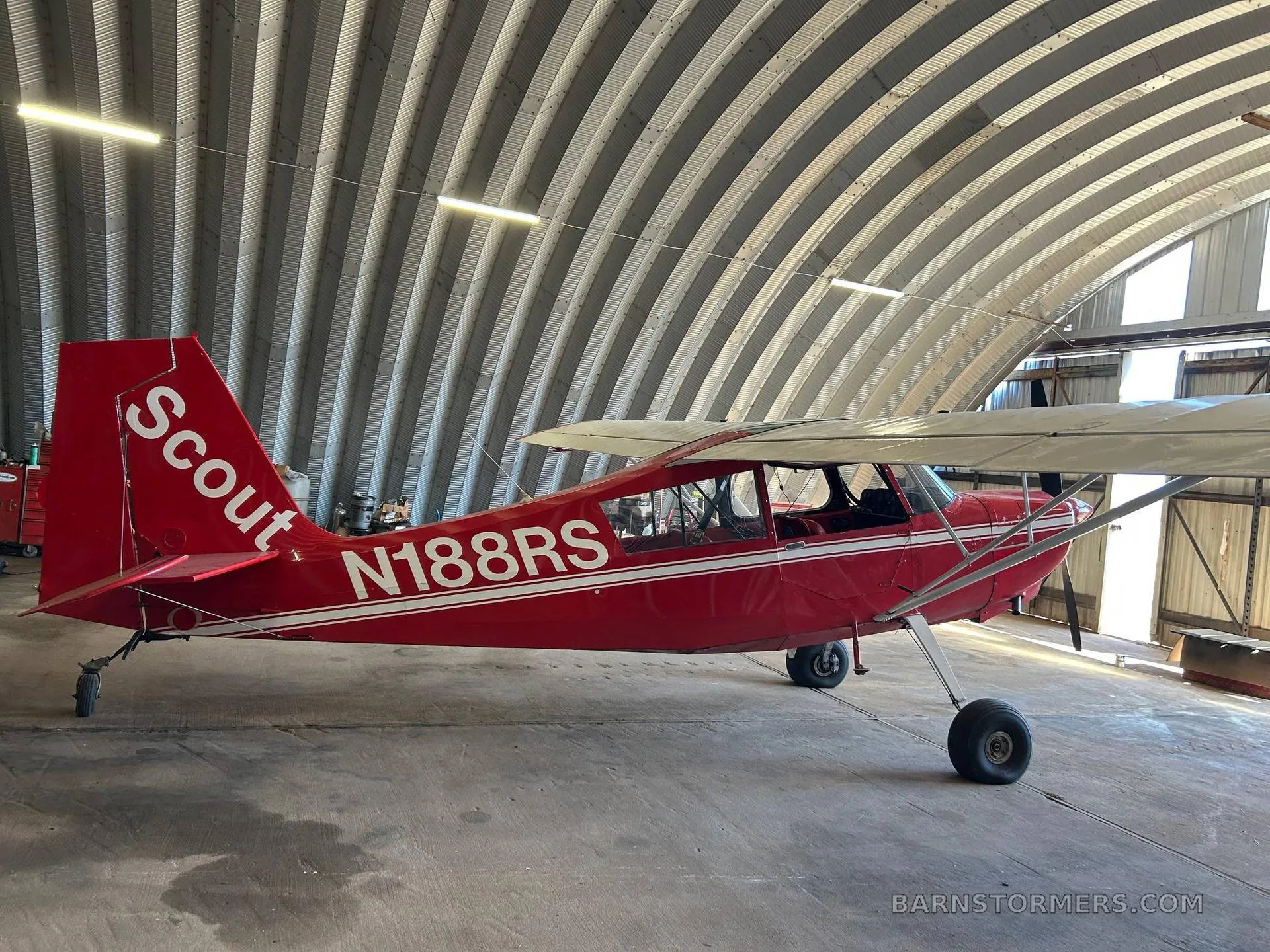 A red Scout N188RS aircraft with white stripes is parked inside a metal hangar, viewed from the rear-left.