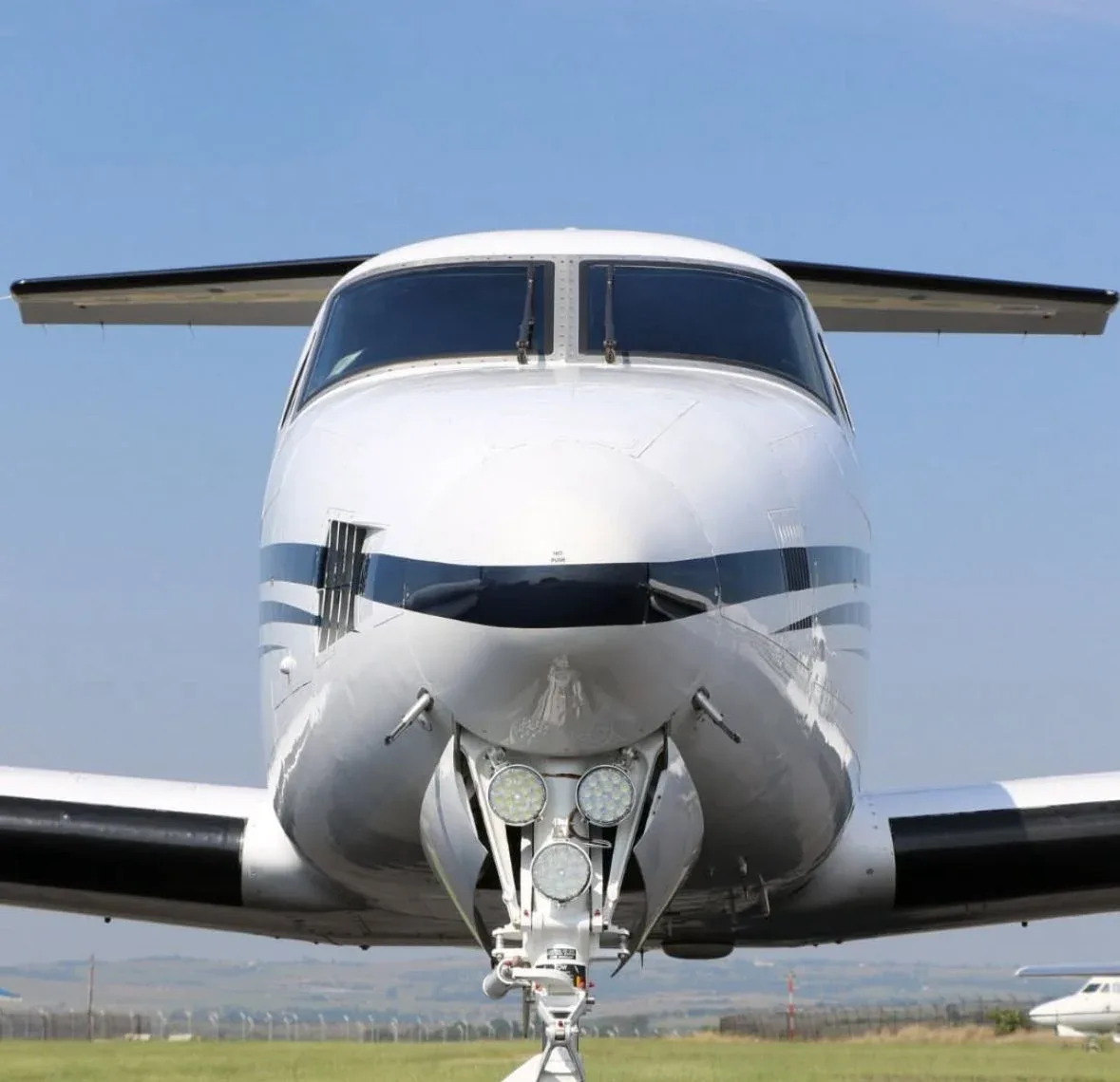 A frontal view of a white business jet with dark stripes on its fuselage, parked on a grassy airfield under a clear blue sky.