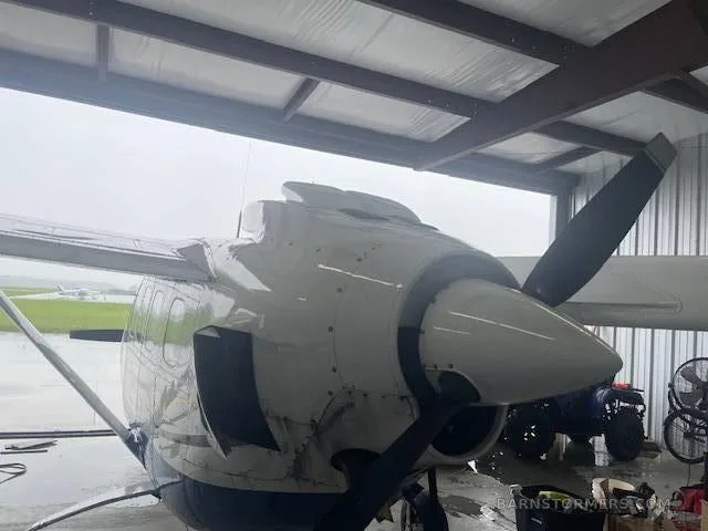 The front engine and propeller of a white twin-engine aircraft, likely a Cessna 337 Skymaster, inside a hangar on a rainy day.