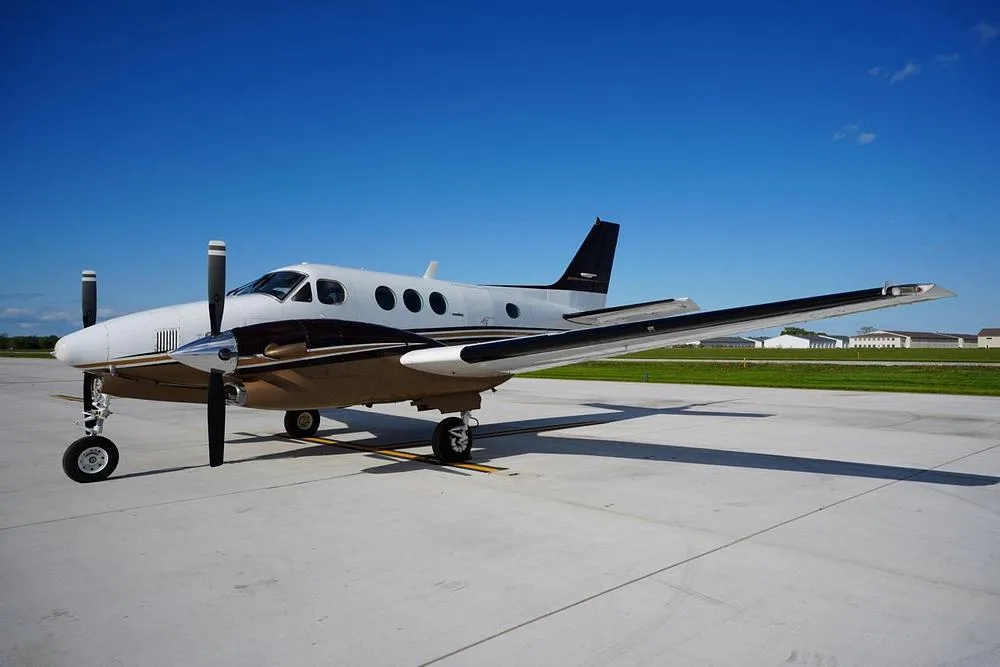 A white and brown Beechcraft King Air 200 parked on an airfield under a clear blue sky.