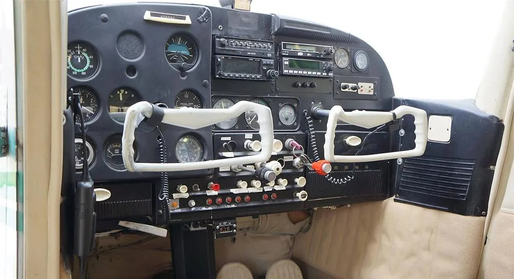 The black instrument panel of a small aircraft, featuring various gauges, radios, and white control yokes.