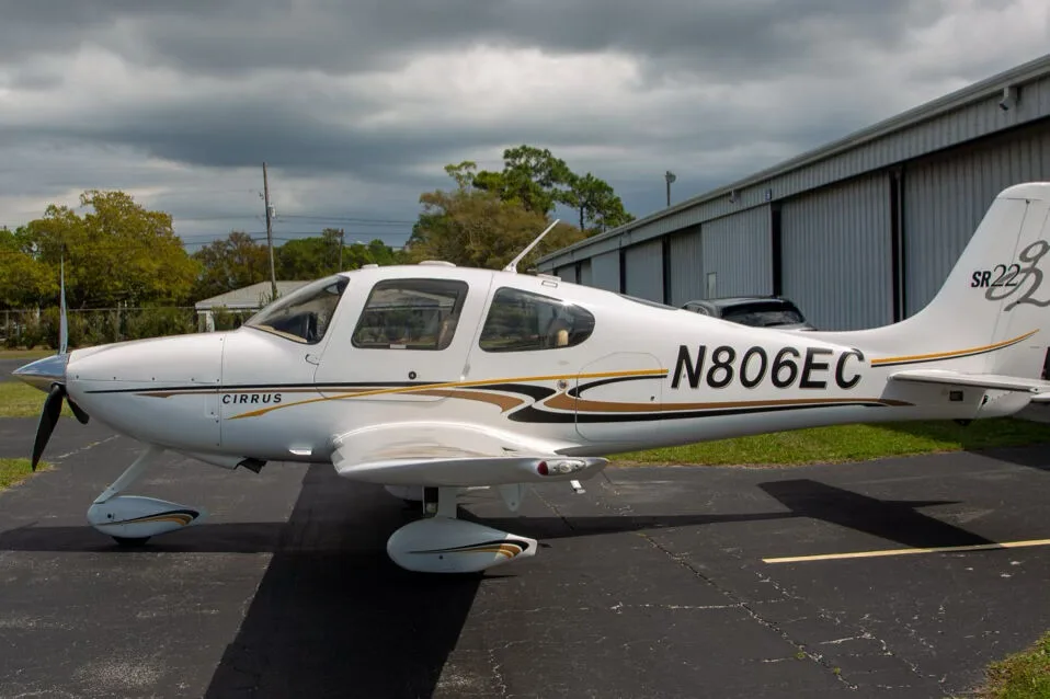 A white Cirrus SR22 aircraft with black and gold stripes, registration N806EC, parked on an asphalt tarmac next to a hangar.